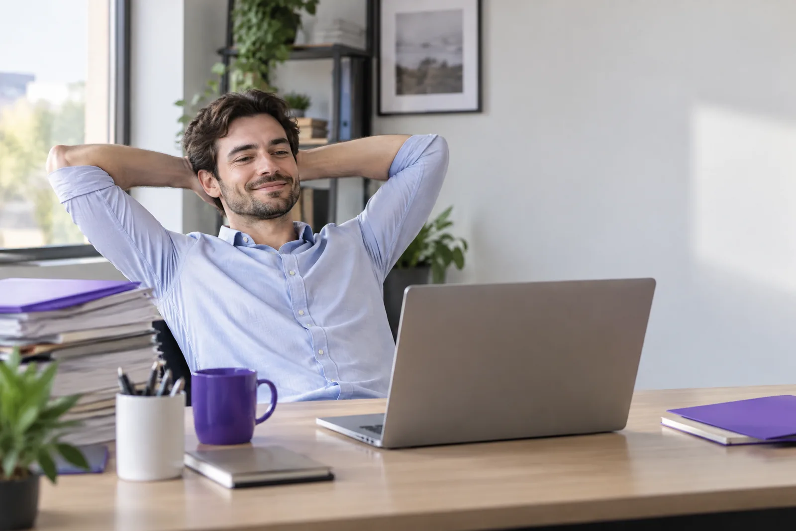 An employee leans back relaxed in his office chair with his hands behind his head, smiling at his laptop; a tall stack of paper sits on the left of the desk.