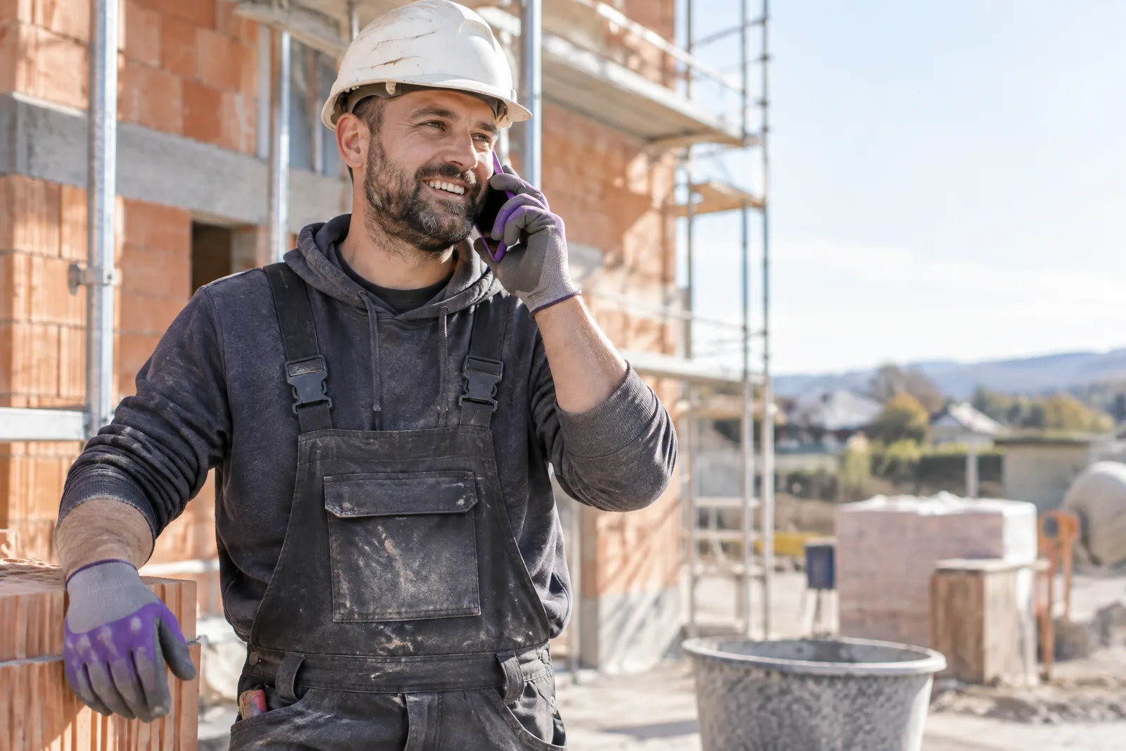 A smiling tradesman in a hard hat and dungarees talks on his mobile phone at a building site in front of a shell structure and scaffolding.